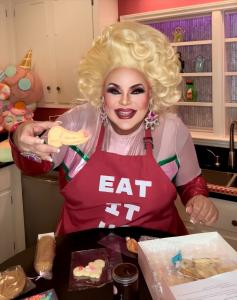 Drag queen Buff Faye, wearing a red “EAT IT UP” apron and glamorous blonde wig, smiles brightly while holding up a decorated penis-shaped cookie from Sugar Wood’s Naughty Cookie Decorating Party kit, showcasing her Big Drag Energy for bachelorette party f