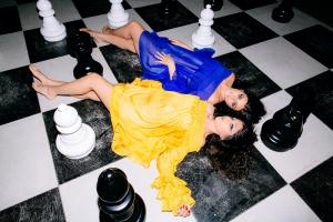 Farrah and Tamara Mechael lie side by side on a black-and-whote chessboard, surrounded by large chess pieces. One wears a bright yellow dress and the other a bold blue dress, symbolizing creativity, balance, and vision in Scorpio Productions’ approach to 