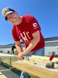 Smiling volunteer in red SHP t-shirt and cap using a hand planer on wooden bed frame components outdoors under clear blue sky.