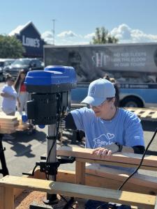 Volunteer in a blue service t-shirt and light blue cap operating a blue drill press on wooden bed frame pieces at an outdoor bed-building event, with SHP trailer and Lowe’s store facade visible in background.