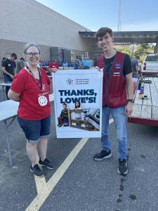 Sleep in Heavenly Peace volunteer in red SHP t-shirt and young Lowe's employee in red vest holding a "Thanks, Lowe's!" appreciation sign featuring the SHP logo and photos of children with bunk beds, standing in Lowe's parking lot with bed-building activit