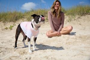 A dog wearing a pink BarkUV sun-protective vest sitting outdoors in the sun with a woman wearing BloqUV enjoying the beach to highlight pet sun safety and adoption awareness.