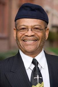 Rev. Dr. J. Alfred Smith, Sr. smiling wearing black suit, white shirt, hat, cross pendant