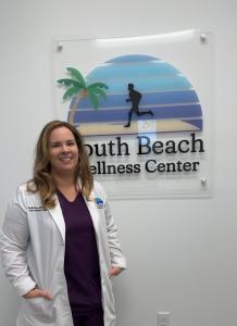 Healthcare provider in a white coat standing beside the South Beach Wellness Center logo sign at the clinic’s Lutz/Wesley Chapel office