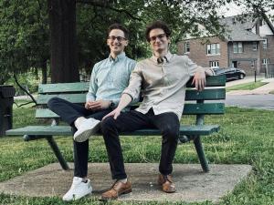 Two young men sit together on a green wooden bench in a park. Both are smiling at the camera; one wears a light blue button-down shirt with dark pants and white sneakers, while the other wears a beige button-down shirt with dark pants and brown shoes. Tre