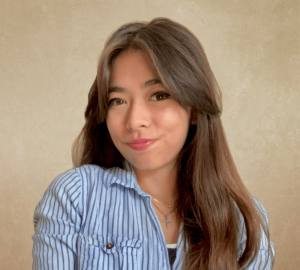 Smiling woman with long brown hair wearing a blue striped shirt and a necklace, against a beige background.