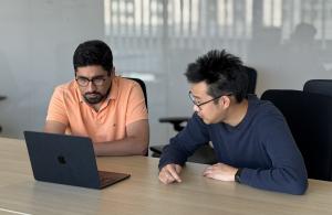 Two male researchers sitting on the same side of a table staring at a black laptop computer