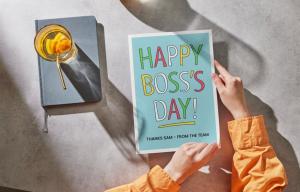Hands holding a colorful “Happy Boss’s Day” card on a desk with a notebook and drink, symbolizing workplace appreciation.