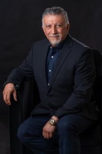 Hamid Kohan, CEO of Legal Soft, seated in a dark suit with a patterned navy shirt and gold wristwatch, smiling confidently against a dark background.