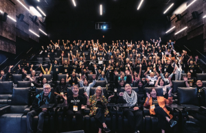 Large group photo inside a theater with audience members smiling and raising their arms, seated behind artist Nobuko Miyamoto and filmmaker Tadashi Nakamura in the front row during the 2024 Silicon Valley Asian Pacific Film Festival.