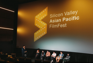 Moderator Mike Inouye, NBC Bay Area weekday morning traffic anchor, stands with a microphone in front of a large movie screen displaying the Silicon Valley Asian Pacific FilmFest logo, as six filmmakers sit on stage for a panel discussion.