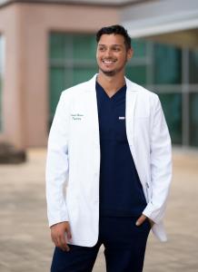 Smiling male healthcare provider wearing white medical coat over navy blue scrubs standing outdoors in front of medical facility