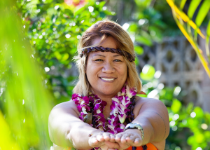 Smiling Pacific Islander woman wearing purple orchid lei and traditional headband outdoors in Hawaii with lush green foliage in background
