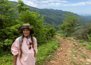 Young woman with braided hair wearing pink shirt and hiking hat standing on mountain trail with green hills in background