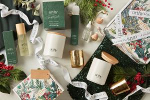 A festive flat lay featuring Urthy’s holiday collection, including white candles with wooden lids, a gold votive, room sprays, oils, and decorative gift boxes with greenery, pinecones, and red berries.