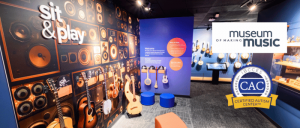 Interactive music exhibit featuring guitars and speakers, with signage for the Museum of Making Music and Certified Autism Center badge.