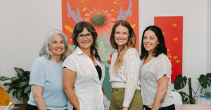 Vervain and Good Apple leaders standing together in the apothecary workspace, smiling before a vivid red floral mural and indoor plants