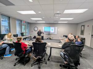 Tanya H. Miller, founder of Secret Weapon Media Agency, stands at the front of a conference room presenting a slide to a group of attendees. She gestures toward the projection screen while participants seated at tables listen and take notes.