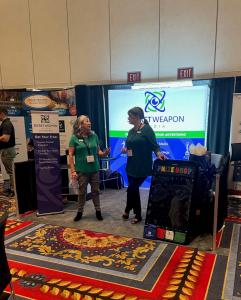 Tanya H. Miller of Secret Weapon Media Agency talks with an attendee in front of the company’s branded trade show booth, featuring the Secret Weapon Media logo, tagline, and service offerings on a large display backdrop.