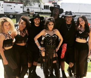 Farrah Mechael stands center stage with her dance team outdoors in coordinated black outfits. Farrah wears a black embellished corset and fringe skirt, posing confidently with hands on hips, symbolizing her performance momentum and growing fanbase.