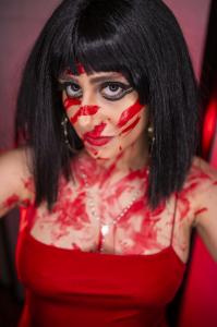 Farrah Mechael poses in a red dress with bold black eyeliner and streaks of red paint smeared across her face, chest, and shoulders. Her piercing gaze under harsh red lighting intensifies the dark and horror-inspired atmosphere of the “Nightmare” visuals.