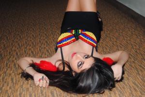 ALT TEXT: Farrah Mechael lies on the floor in a black skirt and colorful beaded top with red feather accents. Her bold eyeliner, long lashes, and red lipstick add a dramatic contrast, bringing out the surreal, carnival-inspired aesthetic of “Nightmare.”