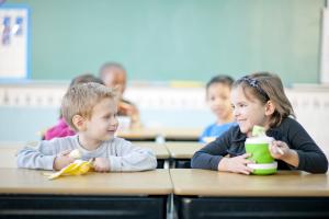 Two smiling children sitting at classroom desks, sharing lunch with other students in the background, representing the focus on creating safer school environments for kids at risk of severe allergic reactions.