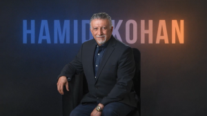Portrait of Hamid Kohan, CEO of Legal Soft, seated in a black chair wearing a dark suit with a patterned navy shirt and gold watch, smiling at the camera. Behind him, the name ‘Hamid Kohan’ is displayed in large gradient letters across a dark background, 