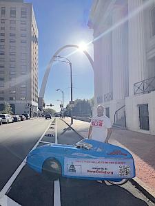 Aaron Novinger standing in front of the Gateway Arch in St. Louis next to his bright blue velomobile during his 2,000-mile charity ride, “Pedaling Against Ponzi’s,” to support victims of financial fraud.