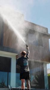 A professional exterior cleaner using a high-pressure washer to clean the upper level of a modern home on the Gold Coast under clear blue skies.