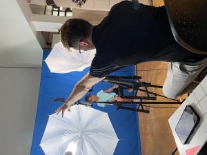 An acting coach gives direction to a young actor seated in front of a blue backdrop during a filming session. The setup includes studio lights with white umbrellas, a camera on a tripod, and a tablet on a nearby table. The coach is gesturing with his arm 