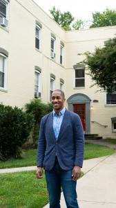 Chris Grant in a deep blue blazer, light blue button down shirt and no tie, wearing dark denim jeans smiles as he stands in front of his company's latest real estate development, the polished beige building and striking red door of Riverdale Station Apart