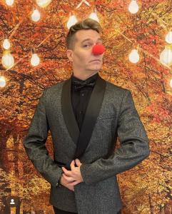 John Stevens, acting coach, poses humorously with a bright red clown nose, wearing a textured grey jacket and black bow tie against an autumn-themed backdrop with vibrant leaves and hanging lights.