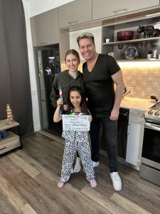 In a modern kitchen, acting coach John Stevens, dressed in a black V-neck T-shirt and jeans with blonde highlights, stands with a smile next to Kate Roman from The Next Step. She is on the left, with light brown hair tied back, wearing a dark green hoodie
