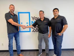 Billy Cline, CEO of YourNewDoor.com, shakes hands with the former owner and sales manager of Dallas Door Designs to commemorate the acquisition. All three men are smiling and dressed in branded company apparel, marking the beginning of their new partnersh