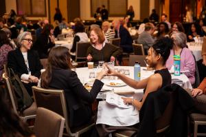 A lively group of people sit at round tables in a conference room, smiling and chatting. Two women in the foreground laugh and touch fingertips in a playful gesture while others around them converse and enjoy refreshments.