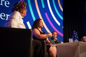 Three panelists sit on stage during a conference, speaking and listening intently. One woman in the center gestures while talking, with colorful graphics displayed on the screen behind them and water bottles on a table in the foreground.