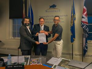 Three men stand together holding the signed resolution on Hinduphobia at Wainwright, Alberta. From right to left: Wainwright resident Roshan Prasad, Wainwright Mayor Bruce Pugh, and CoHNA Canada president Rishabh Saraswat. Four flags and the Wainwright to