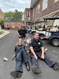A behind the scenes photo of the director and stars sharing a moment of laughter. The director Carol Ray Hartsell (50, white) stands over Sean (40's white) and Moiya (30, black) who lie stretched out, exhausted, on a small road of the movie lot lined with