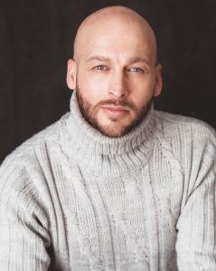 The image shows a bald man with a well-groomed beard, wearing a light gray cable-knit turtleneck sweater. He's looking directly at the camera with a neutral expression. The dark background contrasts with his sweater, emphasizing his face. The soft lightin