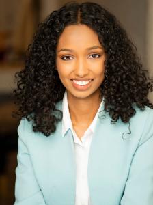 Professional headshot of a young woman with curly black hair, smiling confidently in a light blue blazer and white blouse against a softly blurred indoor background.