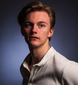 The image shows a young man with short light brown hair, wearing an off-white, textured long-sleeve polo shirt featuring a subtle checkered pattern. He has an open button at the neckline and is gazing slightly upward with a thoughtful expression. Dramatic