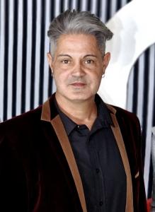 The image shows a person with short, grey hair styled upwards, wearing a velvet jacket with brown lapels over a black collared shirt. The background has vertical black and white stripes. The person has a neutral expression, is facing the camera directly, 