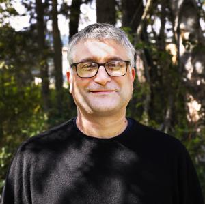 Mountain Rose Herb's Executive Vice President, Steven Yeager. Image feature headshot of Steven smiling in a black t-shirt and black eyeglasses behind a wall of trees.