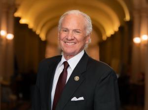Governor Henry D. McMaster smiling in a dark suit and burgundy tie, standing in a warmly lit hallway. He has white hair and is wearing a lapel pin and pocket square.