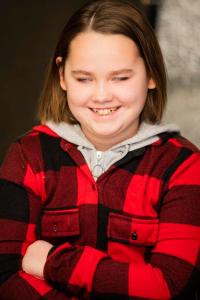 A person with short brown hair, wearing a red and black checkered jacket over a gray zip-up hoodie, smiles with their eyes closed. Their arms are crossed and the background is softly blurred, with warm lighting giving the scene a cozy and inviting feel.