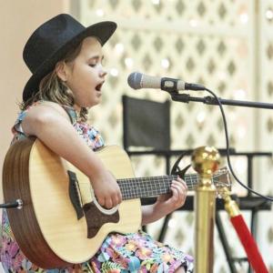 ThA young girl in a black hat and colorful floral dress sings while playing an acoustic guitar. She sits in front of a microphone, immersed in her performance. The background features a chair and a decorative lattice wall with small lights, suggesting a m