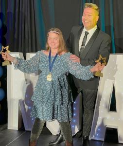 A young woman in a blue floral dress holds two star-shaped trophies and smiles on a red carpet. She wears a headband, gold medal, sparkly leggings, and brown boots. A man in a black suit stands behind her, smiling, with his arm around her shoulder, also h