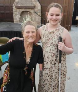 A beautiful woman who is a four way amputee, wearing a formal black dress, stand next to a young girl in a floral dress holding a cane to help her as she is blind. They are in a ballroom at an event.