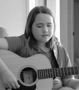 A black and white photo of a young girl playing guitar, wearing a T-shirt.
